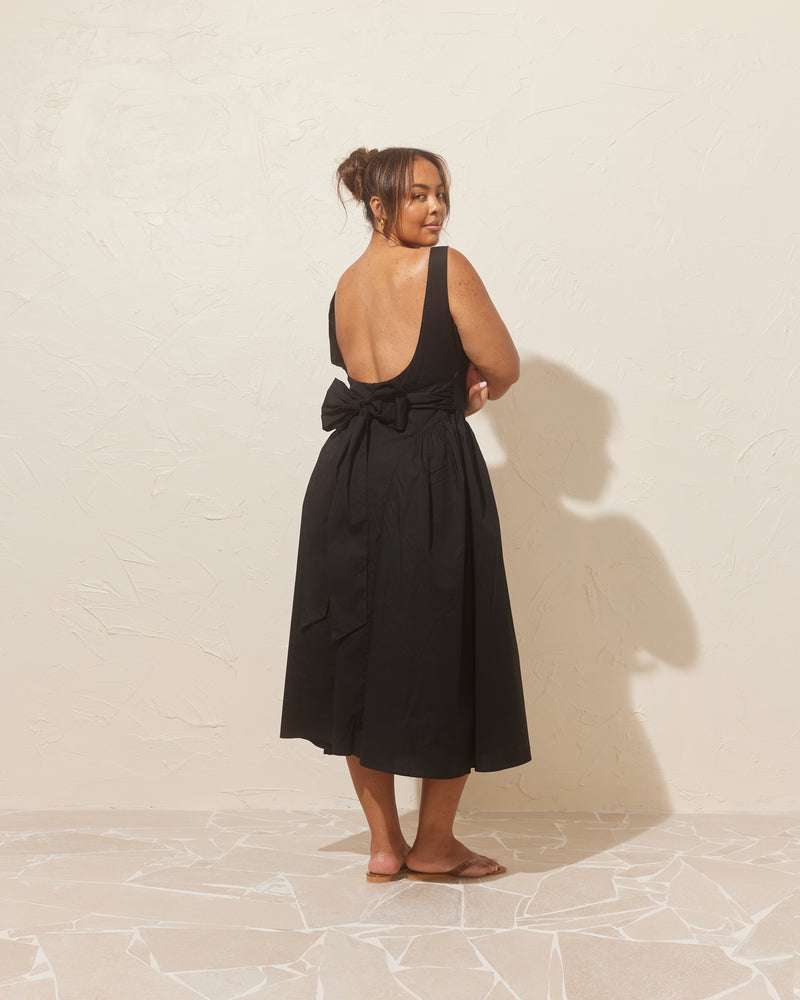 CARMELA BOW DRESS BLACK | Sleeveless midi dress with a high, boat neckline, designed in an organic cotton. This dress has a floaty A-line skirt and turns to reveal an oversized bow at the back.